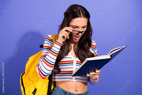 Young female student reads a book with glasses and a yellow backpack against a purple background