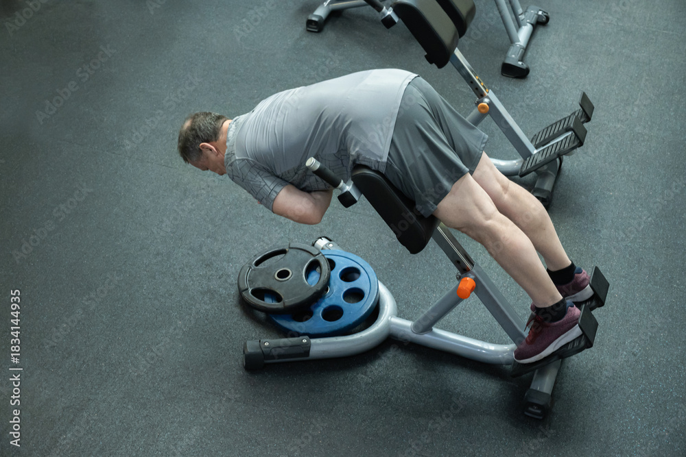 Naklejka premium Adult man performs back extension on bench in gym, strengthening lower back muscles and core stability during workout.