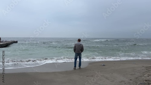 Man standing alone on sandy beach looking at rough sea. Overcast cold day Black Sea Odessa Ukraine. Contemplation, solitude, relax, rest during vacations.