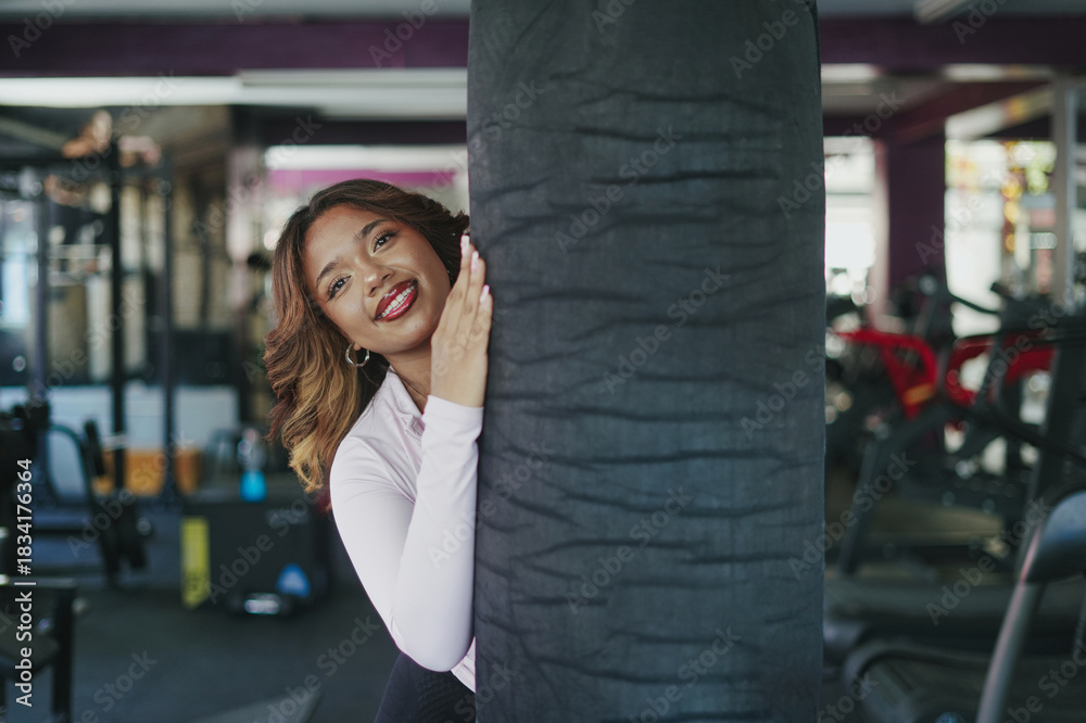 Fototapeta premium Young woman smiling peeking from behind punching bag in gym
