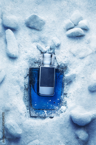 A bottle of fragrance is placed inside ice and snow. The setting shows a mix of icy surfaces and natural stones. The scene captures a moment of cold beauty.