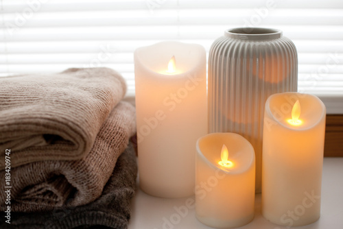 Pile of warm knitted sweaters, led battery candles and vase over white background with soft window light.