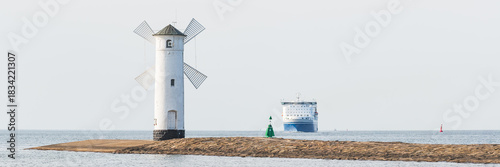 Stawa Mlyny, a tourist attraction in Swinoujscie, a windmill-shaped navigation sign stands at the end of the breakwater at the mouth of the sea, a large ferry on the horizon.