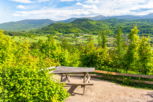 Fototapeta Naklejka Na Ścianę i Meble -  The Giant Mountains, Western Sudetes, with a rest area on a mountain hiking trail overlooking a mountain valley and mountains with dense forests on the slopes. A panoramic view on a sunny summer day.