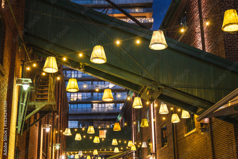 Obraz premium Hanging decorative lamps and warm string lights between brick buildings in Torontos Distillery District at night.