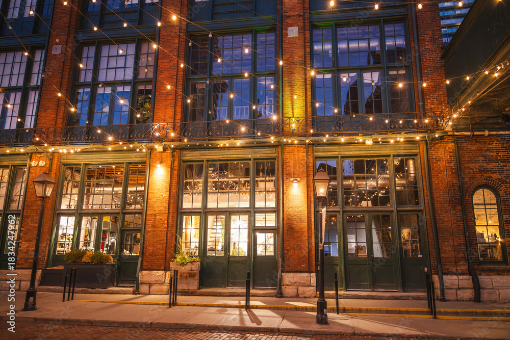Fototapeta premium Evening view of the historic Distillery District in Toronto with illuminated brick architecture, tall windows, and festive string lights.