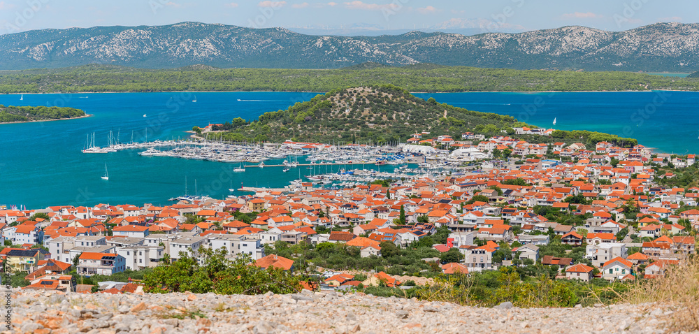 Fototapeta premium The coastal town of Murter in Croatia is situated on the seashore with a mountain landscape, seen from the top of Mount Raduc on a sunny summer day.