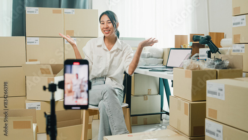 Youth Asia lady owner small business online shop sit front of phone look at camera live streaming talk to customer order promotion with full carton box at home. Shipping and delivery concept.
