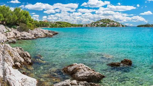 A natural rocky coastline with clear blue waters. A view of the small coastal town of Tribunj on a sunny day, with green trees lining the shore.