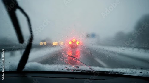 car windshield with moving wipers during heavy snow storm. blurred road view with red brake lights through wet glass. dangerous driving conditions and poor visibility concept. bad weather