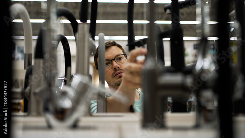 Young man wearing glasses carefully examining various faucets displayed in a hardware store, considering different models before making a purchase