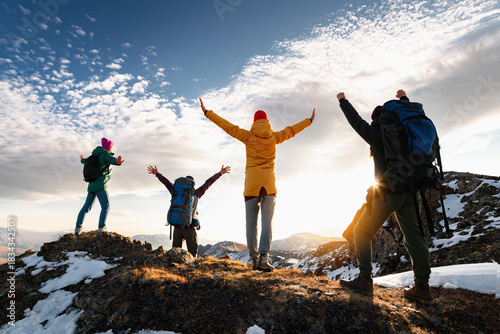 Group of happy young adult friends hikers or tourists with backpacks are standing in winner poses with open arms at mountain top and looks at sunset