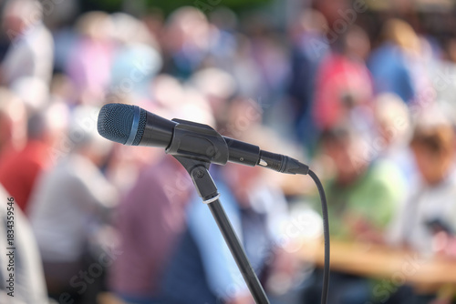 Seitliche Nahaufnahme eines Mikrofons im Halter vor einer Masse an unkenntlichen Menschen im Publikum im Hintergrund auf einer Open-Air Veranstaltung – selektiver Fokus, sehr viel Tiefenunschärfe