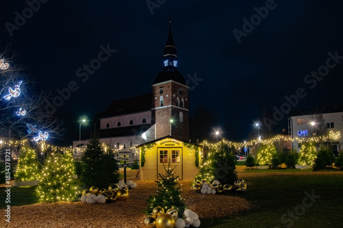 Festive Christmas lights, decorated trees, and a small holiday house glow warmly in front of a historic church on a winter night.
