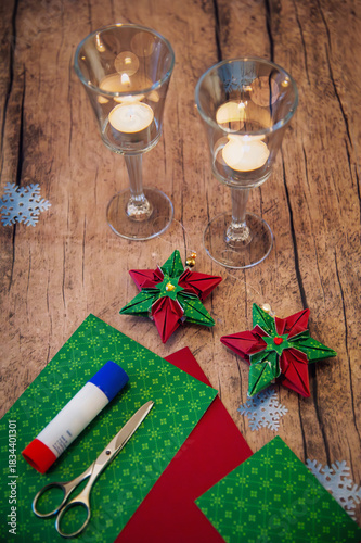 Christmas crafts at home: red and green 6-pointed paper stars with scissors and glue stick on a table in front of festive candlelight in vertical format.