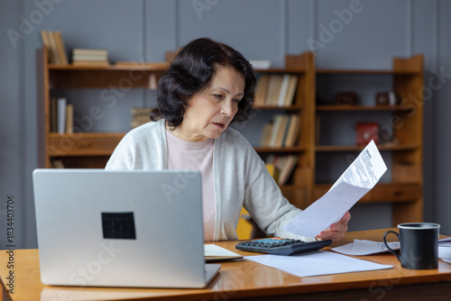 Middle aged senior woman sit with laptop and paper document. Pensive older mature lady reading paper bill pay online at home managing bank finances calculating taxes planning loan debt pension payment