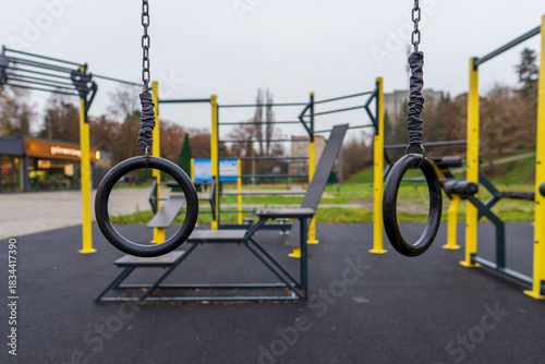 Fotografie Empty black fitness rings hang suspended at an outdoor public gym, featuring modern yellow and black exercise equipment for calisthenics training