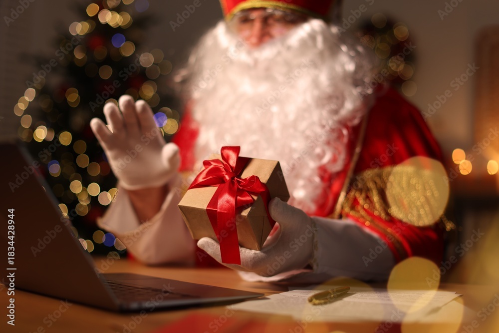 Obraz premium Saint Nicholas with gift and laptop at desk in room decorated for Christmas, selective focus