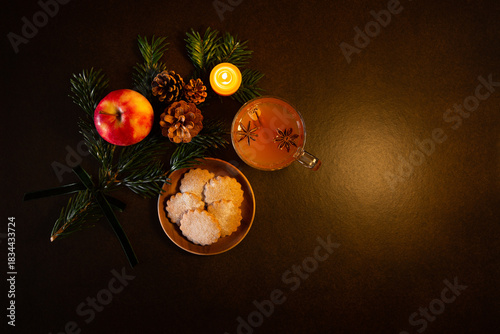 Décor de Noël, branche de sapin vert, pomme, pommes de pin, bougie en cire naturelle allumée, petits gâteaux, tasse en verre avec jus de pomme chaud et épices de Noël, vue du dessus, fond brun foncé