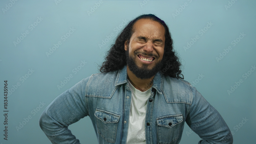 Fototapeta premium Man wearing denim jacket with hands on hips looking down in a pale blue studio; frustration fatigue.