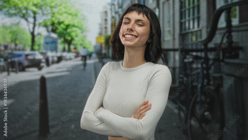 Fototapeta premium Woman wearing white crop top stands smiling with arms crossed on a bustling city street lined with parked bicycles; confidence.