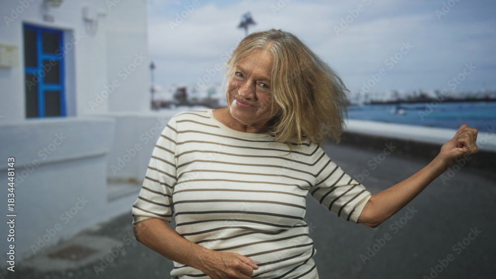 Fototapeta premium Senior woman with raised hand and open arm gesture laughing on a coastal street beside white building and seawall, wearing striped shirt; joy.