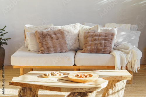Comfortable white sofa with decorative pillows, cushions and wooden coffee table in cozy minimalist living room interior with rustic wooden elements and soft natural light in scandinavian style