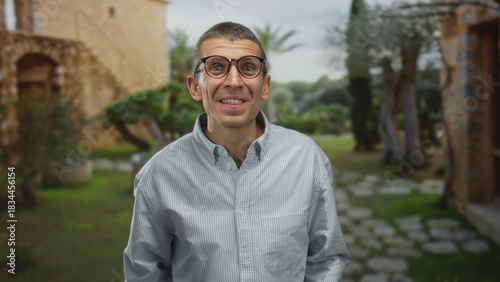 Smiling man with glasses in striped shirt standing outdoors on green pathway near rustic building and trees, expressing surprise and happiness.