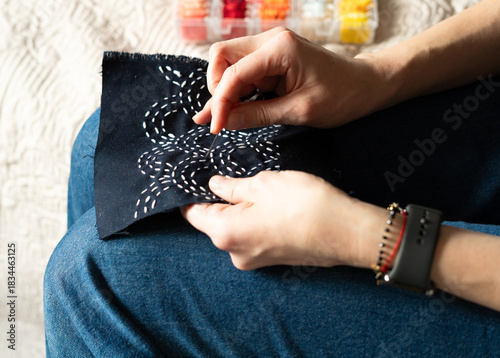 Person does hand embroidery work with a needle on dark fabric while sitting on a chair in a home setting