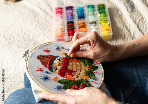 woman works on an embroidery project featuring a bird wearing a hat and scarf. The setting is a home with soft lighting that creates a relaxed atmosphere.