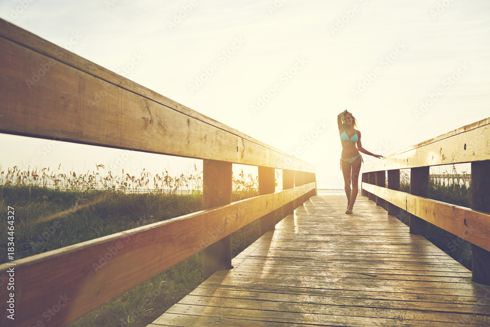 Fototapeta premium Happy woman, bridge or boardwalk at beach with sunset, low angle space and vacation for summer travel. Adventure, portrait or person in bikini for coastal holiday, sunshine and walk on wood pathway