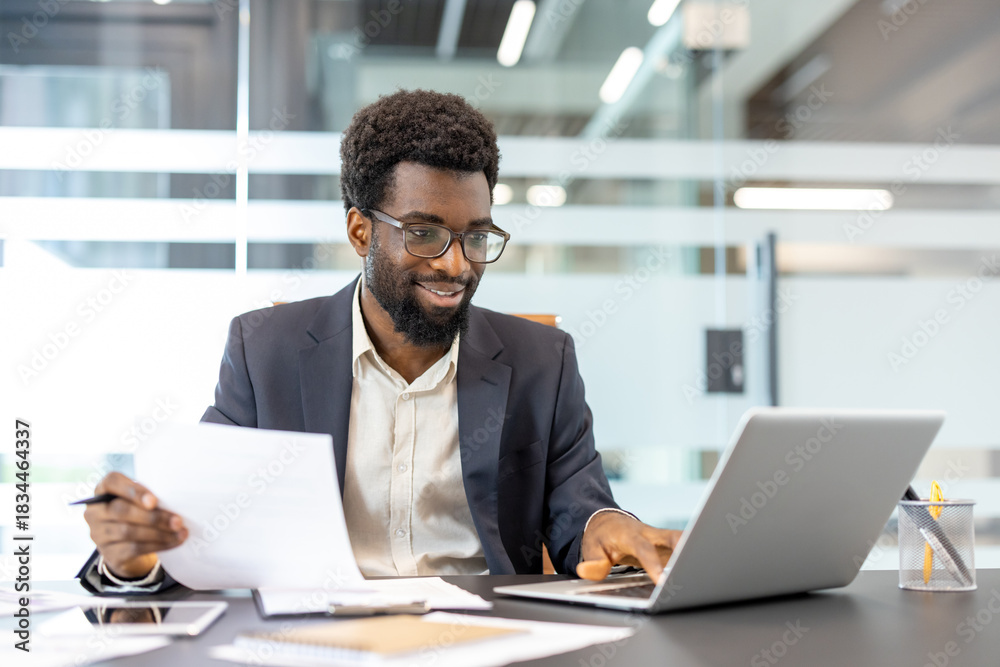 Obraz premium African american businessman wearing glasses smiling and working proactively at an office desk, analyzing documents and typing on a laptop with a modern background