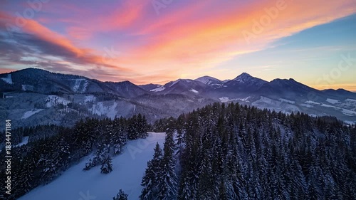 Aerial hyperlapse over winter mountain forest and snowy peaks under colorful evening sunset sky