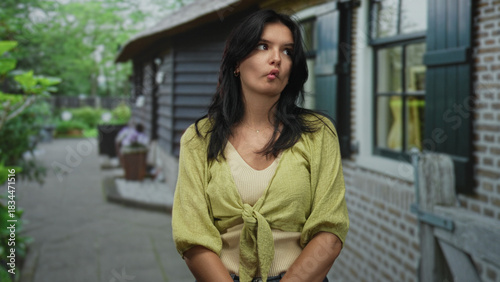 Tableau sur toile Woman wearing green cardigan clasps hands on a leafy street beside a brick building with potted plants; confidence