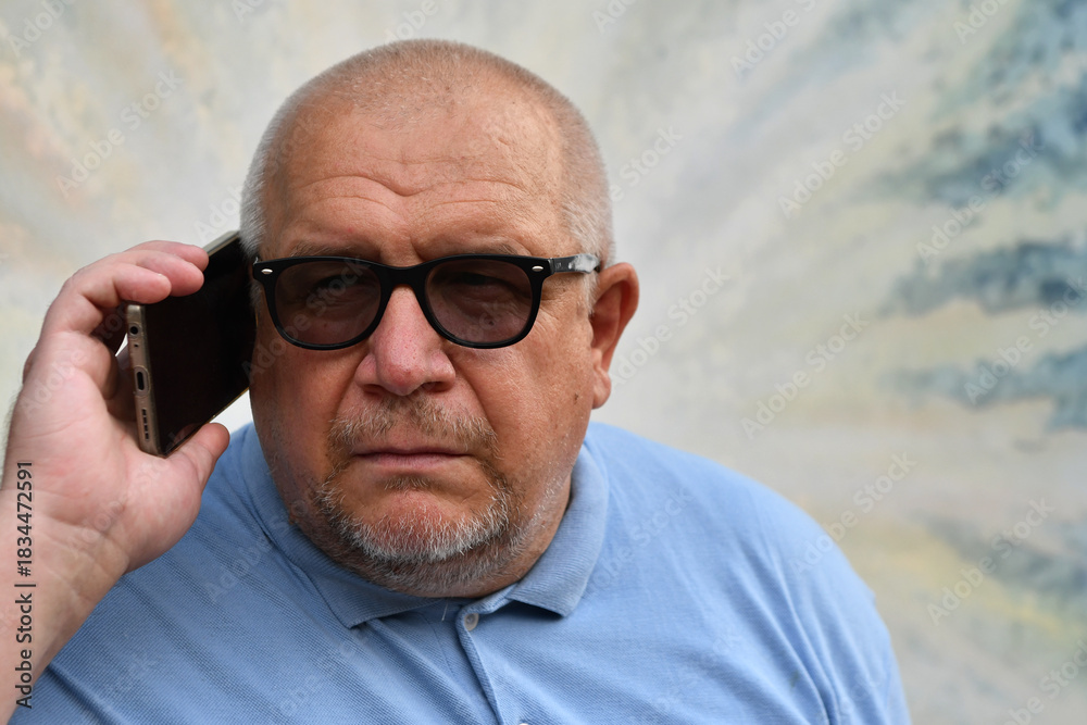Obraz premium An elderly man wearing glasses and a blue polo shirt and glasses holds a phone to his ear.