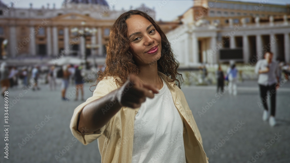 Obraz premium Young african american woman in a beige shirt points finger to camera in st peters square outside the vatican among tourists and colonnades; confidence travel invitation.