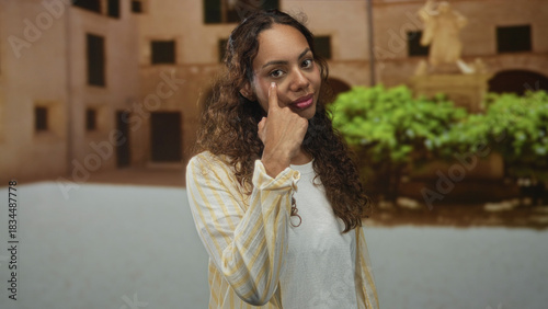 Woman points finger to cheek beside a building in an old town plaza with direct gaze and posed stance; confidence self belief.