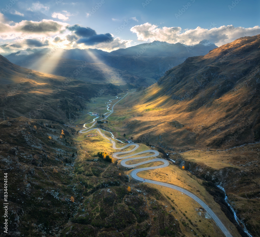 Obraz premium Aerial view of winding mountain road in the Swiss Alps, Switzerland, with sharp curves, golden slopes, valley and dramatic sun rays breaking through clouds over alpine peaks. Top drone view of road
