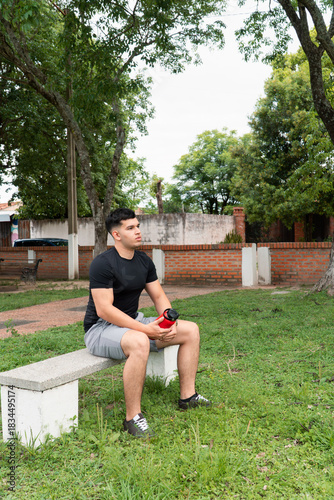 Man holding a red water bottle while sitting at the bench park.