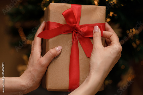 Hands carefully tie a red ribbon around a wrapped Christmas gift, creating a festive presentation