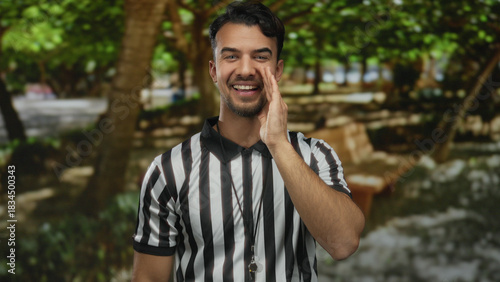 Young hispanic man referee gesturing at outdoor park with lush green trees wearing striped uniform and whistle demonstrating excitement and focus during sports activity.