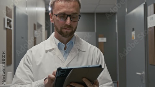 Front view close up shot of professional male doctor holding digital tablet and wearing lab coat while walking towards camera in clinic hall, focus flowing from hands to face, copy space