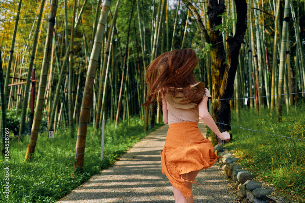 Obraz premium Woman running along a winding bamboo forest path in summer, orange skirt flowing, back view showing motion and carefree movement among tall green stalks on a shaded trail.