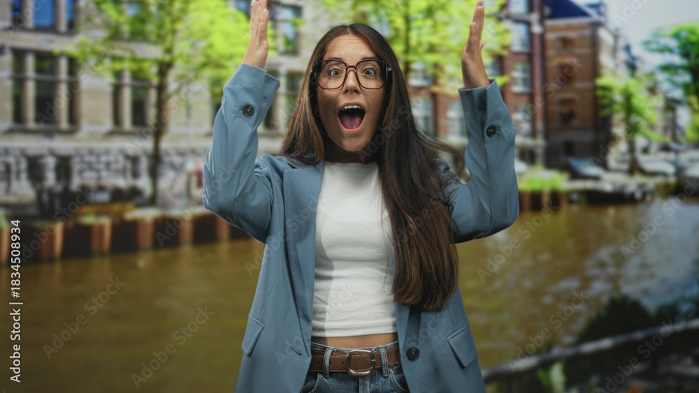 Fototapeta premium Young hispanic woman with hands raised and open mouth in surprise, wearing glasses and a blazer, standing on a street beside a canal; surprise travel.