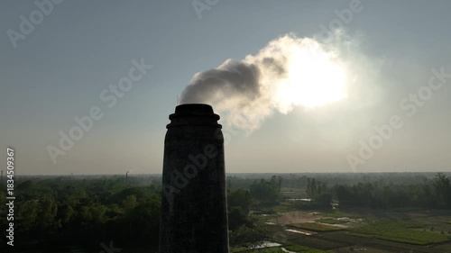 Thick smoke bursts from a brickfield chimney under the blazing sun, filling the sky and quietly harming nearby habitats, showing the harsh impact of industrial pollution.