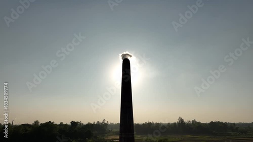 Thick smoke bursts from a brickfield chimney under the blazing sun, filling the sky and quietly harming nearby habitats, showing the harsh impact of industrial pollution.