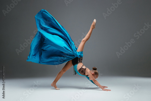 Cheerleader dancing with flying blue cloth fabric. Graceful ballerina dancing on grey studio background. Dancer in sport leotard doing Split. Child, artist, movement, action and motion concept