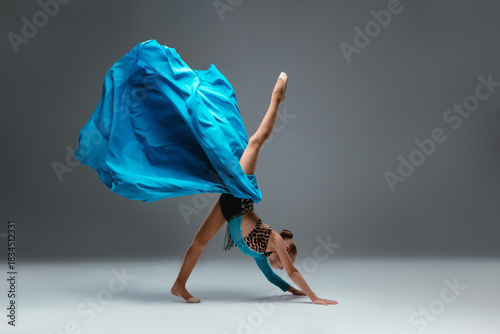 Graceful ballerina dancing on grey studio background. Cheerleader dancing with flying blue cloth fabric. Dancer in sport leotard doing Split. Child, artist, movement, action and motion concept