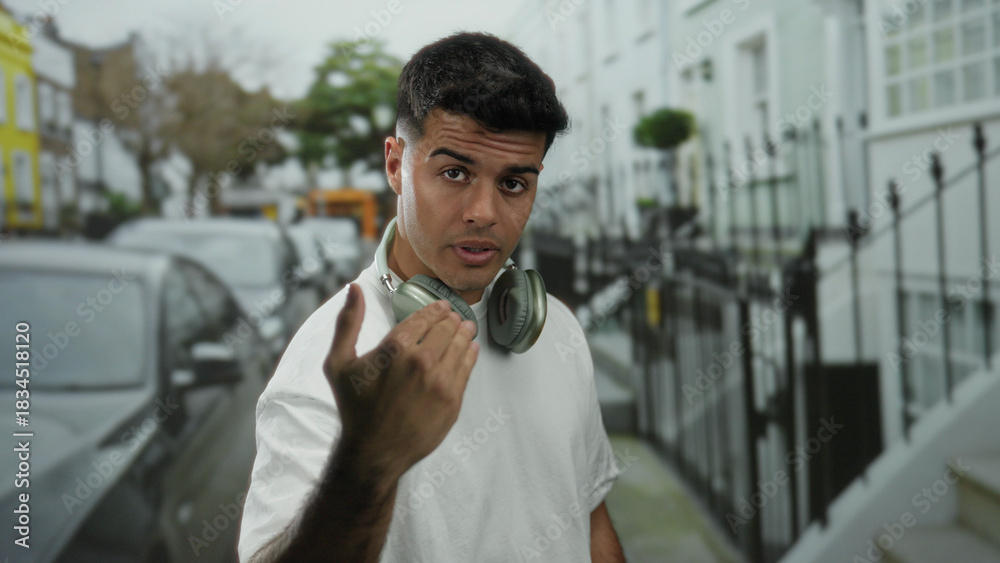 Obraz premium Young man with headphones standing on a city street surrounded by parked cars and buildings.