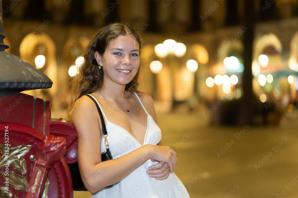 Obraz premium Young woman is walking around Barcelona at night, she stopped near a pole in the square. European woman walking on a summer evening